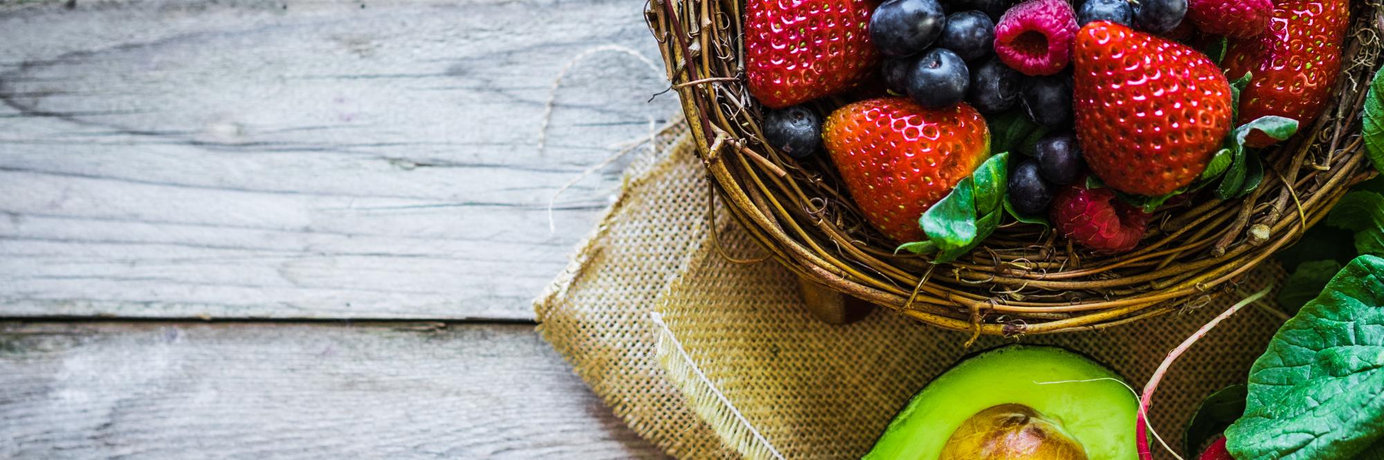 Close-up of a basket with strawberries and blueberries, half an avocado, and a bunch of radishes on a wooden table.